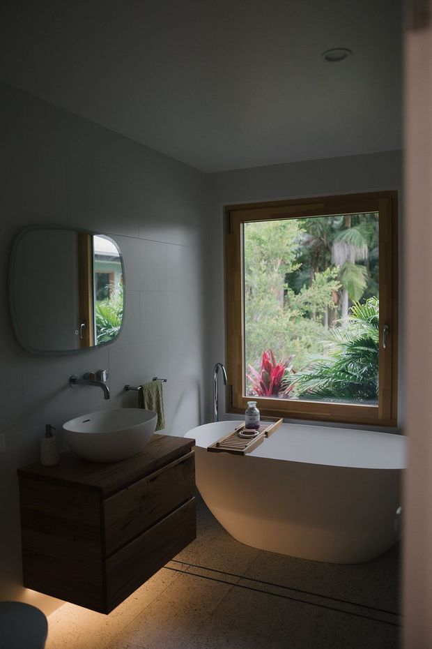 A dimly lit contemporary bathroom featuring a freestanding white bathtub positioned beneath a large timber-framed window, a wall-mounted dark timber vanity with a white vessel basin, wall-mounted tapware, a rounded mirrored cabinet, and warm-toned textured flooring. Lush greenery is visible outside the window.