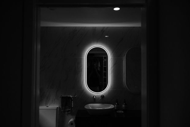A dark, atmospheric bathroom viewed through a doorway. The room features a backlit oval mirror above a vessel basin on dark cabinetry, dark veined wall panels, a toilet, towel rail, recessed ceiling lighting, and subtle black fixtures.