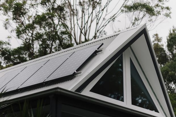 Close-up exterior view of a contemporary gable-roof building, featuring dark cladding, a light metal roof with integrated solar panels, and prominent triangular floor-to-gable glazing. Trees are visible in the background.
