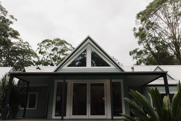 Front exterior view of a dark-clad gable-roof house surrounded by mature trees and landscaping. The facade features a prominent triangular upper window, glazed double entry doors, a broad covered porch or carport-like canopy, and light metal roofing.