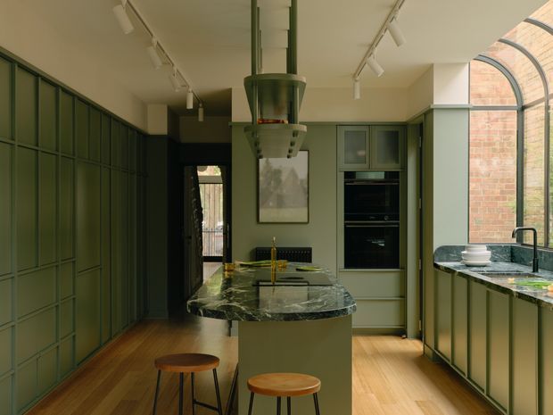 A sophisticated galley-style kitchen with a central island and two timber stools. Deep green floor-to-ceiling cabinetry lines the walls, complemented by matching green cabinetry, dark veined stone benchtops, integrated black appliances, and warm timber flooring. A dramatic sculptural rangehood hangs above the island, while a large arched industrial-style glazed opening brings in natural light and reveals exposed brickwork. Track lighting and restrained artwork complete the refined, moody interior.