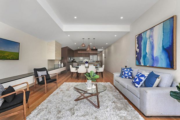 A bright, open-plan contemporary living and dining area with a kitchen in the background. The room features light walls, recessed ceiling lighting, warm timber flooring, a large pale sectional sofa, sculptural glass coffee table, textured light rug, dining table with upholstered chairs, and bold blue abstract artwork and cushions. The space has a clean, airy, refined feel with a long linear layout and integrated cabinetry.