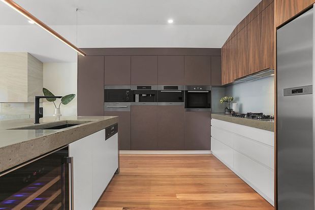 A contemporary galley-style kitchen with a central island, integrated dark cabinetry and appliances, white lower cabinetry, warm timber overhead cabinetry, and a stone benchtop. The room has timber flooring, a pale splashback, recessed ceiling lighting, and a slim linear pendant above the island. A wine fridge is visible beneath the island.