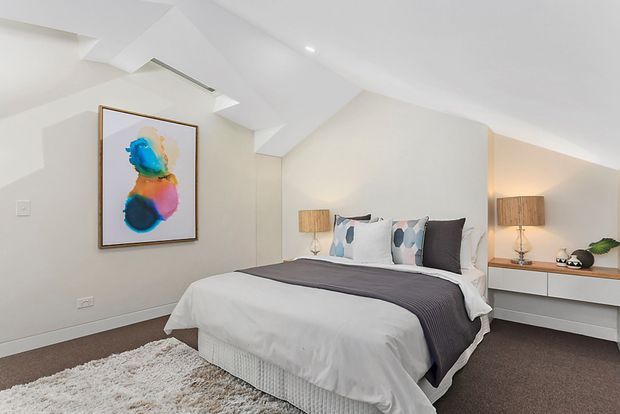 Bright contemporary bedroom beneath a sloped ceiling, featuring a neatly made bed with white bedding and a dark grey throw, patterned accent cushions, integrated white bedside cabinetry, matching lamps, and colourful abstract wall art. The room has soft neutral finishes, recessed lighting, and a plush light rug over dark carpet.