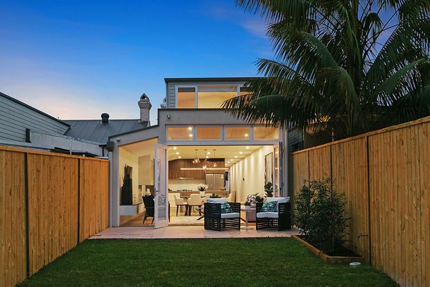 A dusk exterior view of a modern two-storey home extension opening onto a private, fenced backyard. The image is centered on the glazed rear facade and covered entertaining area, with an open-plan living and kitchen space visible inside. Timber privacy fencing, a small lawn, patio paving, warm interior lighting, and tall palm trees create a tropical suburban setting.