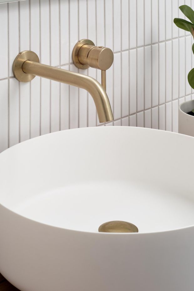 Close-up of a contemporary bathroom featuring a freestanding white bathtub, wall-mounted brushed brass/gold tapware, and narrow vertical white wall tiles. A potted plant is partially visible at the right edge.