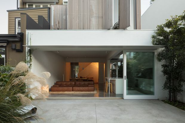 Contemporary multi-storey residential facade with a recessed, open-plan living area opening onto a broad concrete forecourt. The exterior combines white rendered surfaces, vertical ribbed metal cladding and timber-toned siding, with large glazed sliding doors and a sheltered terrace-like opening. Ornamental grasses and a clipped tree frame the entrance, while a brown sofa and warm interior lighting are visible inside.