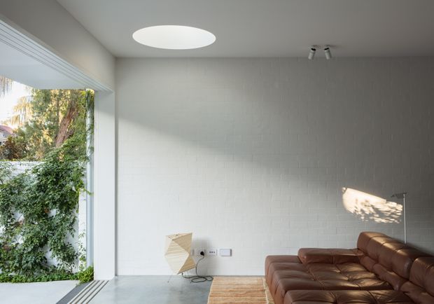 Bright contemporary living room with a large brown leather sectional sofa, pale timber flooring, white-painted brick walls, and a large opening to lush greenery. A circular flush ceiling light, track spotlights, and minimalist furnishings create a clean, airy interior.