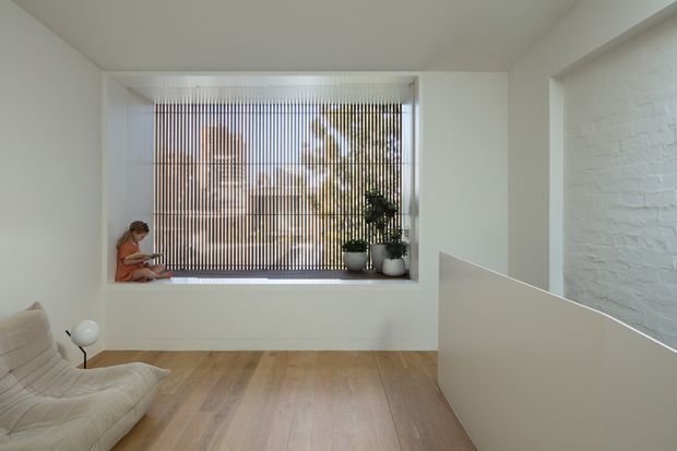 Bright minimalist interior with pale timber flooring, white walls, a white-painted brick feature wall, and a large window fitted with vertical blinds. A low half-height partition separates the main sitting area from an adjacent space, while a neutral lounge chair, floor lamp, plants, and a seated person occupy the foreground and window nook.