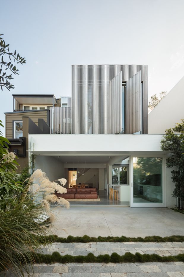 A contemporary multi-level residential facade viewed from the street, featuring a white rendered lower volume, a vertically slatted upper facade with narrow openings, dark timber-toned side cladding, and large glazed openings. A broad concrete forecourt with planted grasses and stone edging leads to an open living area visible through the ground-floor glazing.