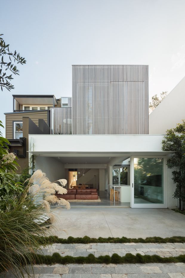 Contemporary multi-storey urban residence viewed from the front, featuring a minimalist white ground-floor facade, broad glazed opening, concrete forecourt, and a distinctive upper-level screen of fine vertical metal battens. Darker timber-toned cladding and projecting volumes appear to the left, while landscaped grasses and clipped planting frame the entry.