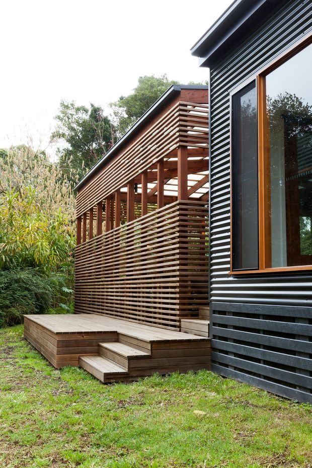 Exterior view of a contemporary residential building in a landscaped setting. The facade combines dark horizontal cladding with warm timber-framed glazing, while a substantial timber pergola or screened deck extends along the side. The deck has horizontal timber privacy slats, built-in stepped platforms, and a small stair descending to the grass.
