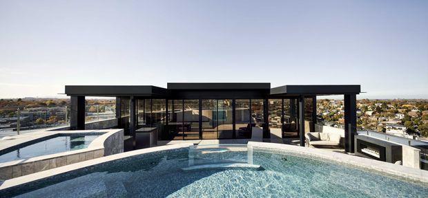 A wide exterior view of a contemporary flat-roof rooftop residence or entertaining pavilion, featuring extensive black-framed glazing, dark structural columns, pale stone or concrete surfaces, and a large curved swimming pool in the foreground. A smaller raised pool or spa is visible to the left, with an outdoor seating and fireplace area to the right. The building overlooks a low-density urban or suburban landscape under a clear sky.
