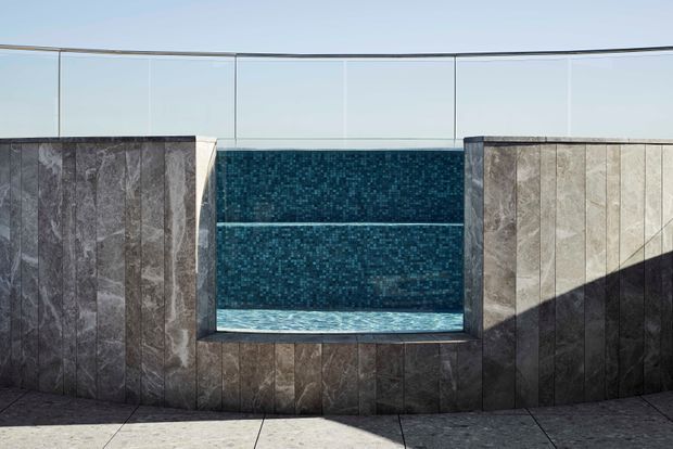 A contemporary rooftop or terrace pool feature with a recessed rectangular pool set between tall stone-clad walls. The pool is finished in dark blue mosaic tile, with a clear glass balustrade along the upper edge and large-format grey paving in the foreground. Strong sunlight creates geometric shadows across the stone surfaces.