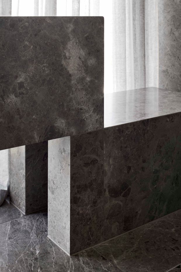 Close-up architectural interior detail featuring a dramatic dark veined stone island or counter with a thick stone benchtop and matching vertical panels. Dark stone flooring continues across the foreground, while sheer floor-to-ceiling curtains provide a softly lit backdrop. The composition is minimalist and material-focused, with no clearly identifiable room function beyond a likely kitchen or reception counter.