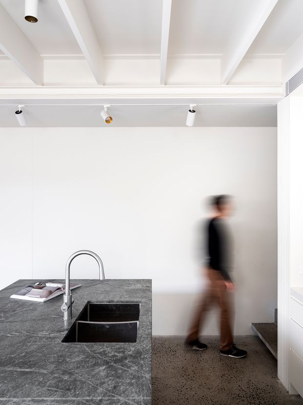 Bright minimalist kitchen interior with a large dark grey stone island, integrated double-bowl black sink and curved chrome tap. White walls and cabinetry are paired with exposed white ceiling beams, track lighting, and a polished concrete or terrazzo-style floor. A blurred person is walking near a short stairway at the rear.