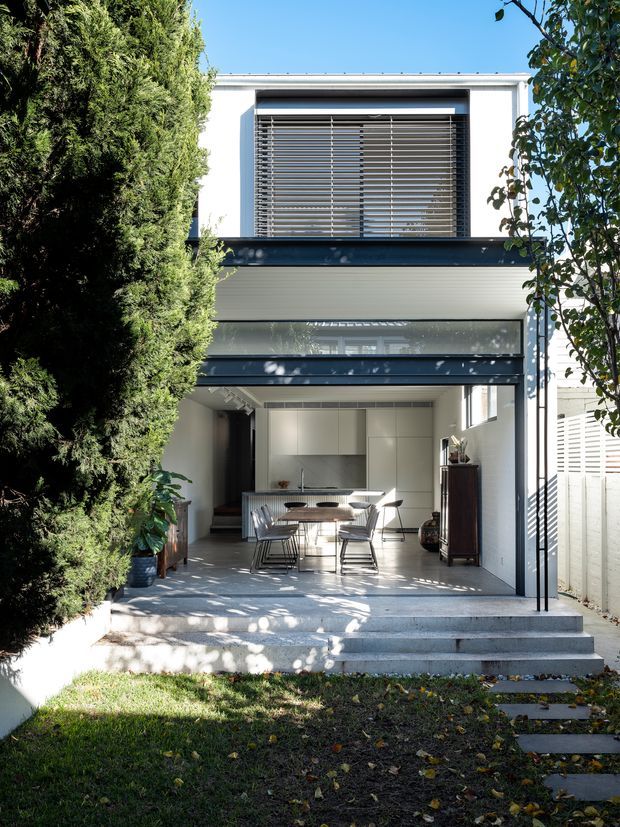 A contemporary two-storey residential rear facade opening onto a small garden. The ground floor has a covered outdoor dining and kitchen area with broad glazed openings, while the upper level features a large window with horizontal external blinds. Clean white walls, dark charcoal framing, pale grey horizontal cladding and concrete steps create a crisp modern composition, partially screened by mature trees.