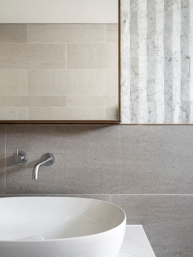Close-up of a refined bathroom vanity featuring a white vessel basin on a pale marble benchtop, wall-mounted chrome tapware, large grey stone-look wall tiles, and a framed mirror. A vertical section of white marble-look mosaic tile provides a contrasting feature behind the mirror.