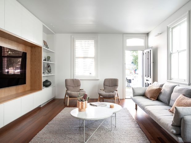 Bright, compact living room with white walls and ceiling, dark timber flooring, a large pale rug, and a built-in white media wall featuring warm timber veneer and open display shelving. A grey sofa, two upholstered lounge chairs, and a round white coffee table create a comfortable seating arrangement. Tall sash-style windows and an open glazed door bring in abundant daylight. The overall design is clean, contemporary, and refined with subtle mid-century-inspired furniture.
