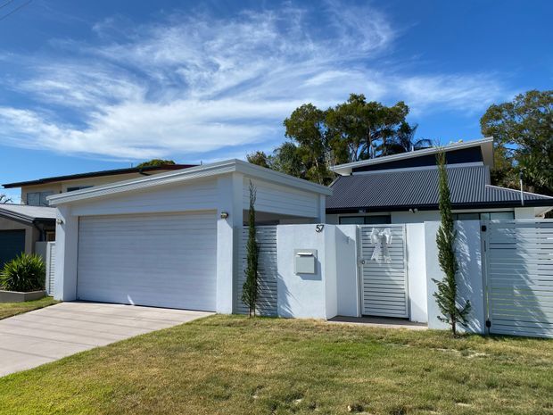 Street-facing view of a contemporary suburban residence with a white double garage, low-pitched dark metal roof, white rendered walls, horizontal slatted screening, pedestrian entry gate, and sparse landscaped planting. The facade has clean geometric lines and a sheltered entry courtyard.