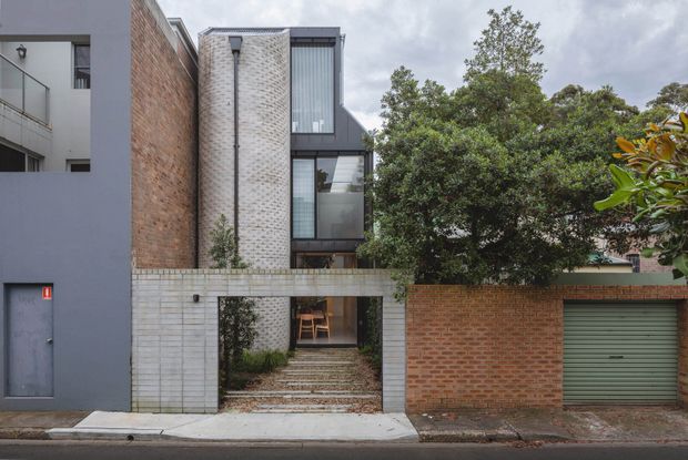 Street-level exterior view of a contemporary multi-storey urban residence or small development. The facade combines red brickwork, pale textured masonry, dark grey rendered walls, and extensive glazed sections. A recessed pedestrian entry passes through a pale masonry wall via a landscaped stepped path, while a red-brick garage frontage with a muted green sectional door occupies the right side. Mature trees partially screen the building.