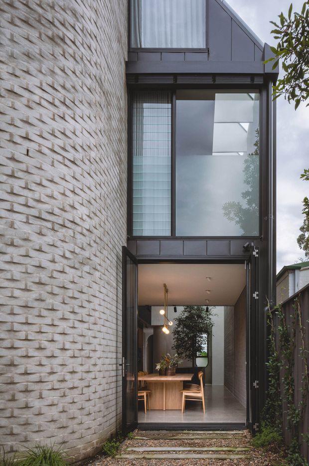 A tall, narrow exterior view of a contemporary residential entry and dining area. The facade combines textured pale brickwork with dark metal-framed glazing and a steep gabled upper section. A large open glass door reveals a timber dining table and chairs, pendant lighting, and indoor planting beyond. Stepping stones lead through a landscaped side passage to the entrance.