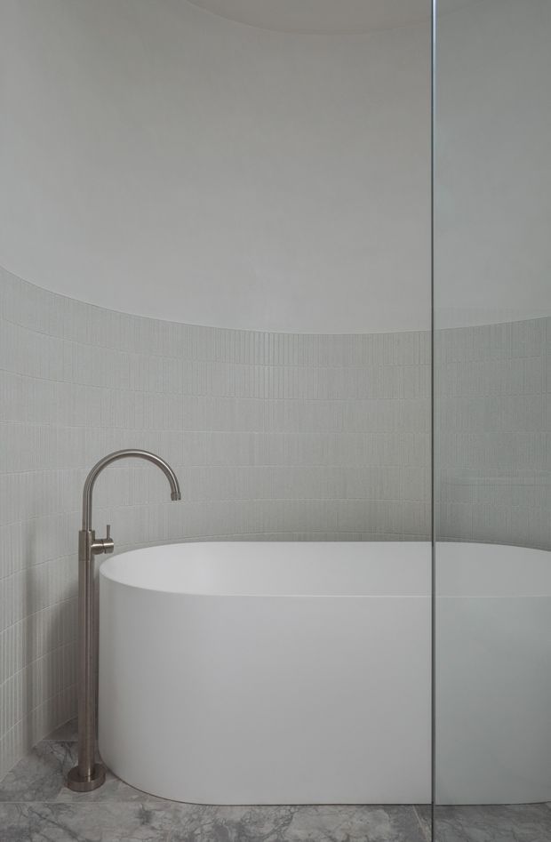 A minimalist bathroom featuring a freestanding white oval bathtub, a brushed metal floor-mounted tub filler, pale vertically textured wall tiles, a curved plaster ceiling, grey marble-look flooring, and a frameless glass shower screen or partition. The restrained palette and curved forms create a serene, refined atmosphere.