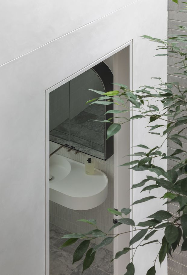 A refined contemporary bathroom viewed through a doorway, featuring a sculptural white curved vanity basin, vertically fluted wall tiles, grey marble-look floor and wall surfaces, and a large arched mirrored cabinet. A leafy plant partially screens the view from the foreground, adding an organic element to the minimalist space.