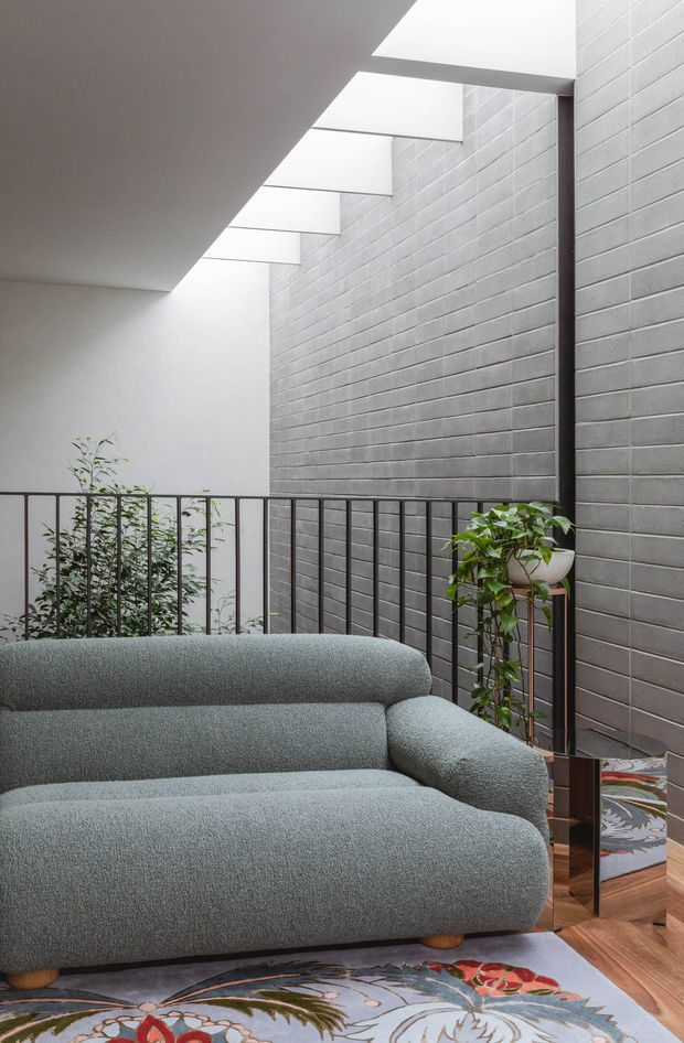 A refined contemporary living area with a large rounded grey upholstered sofa in the foreground, set against a tall light-grey exposed brick wall. A black metal balustrade borders an open internal void or stairwell, with narrow roof-level skylights bringing in soft natural light. Potted greenery, a timber floor, and a patterned area rug add warmth and contrast.