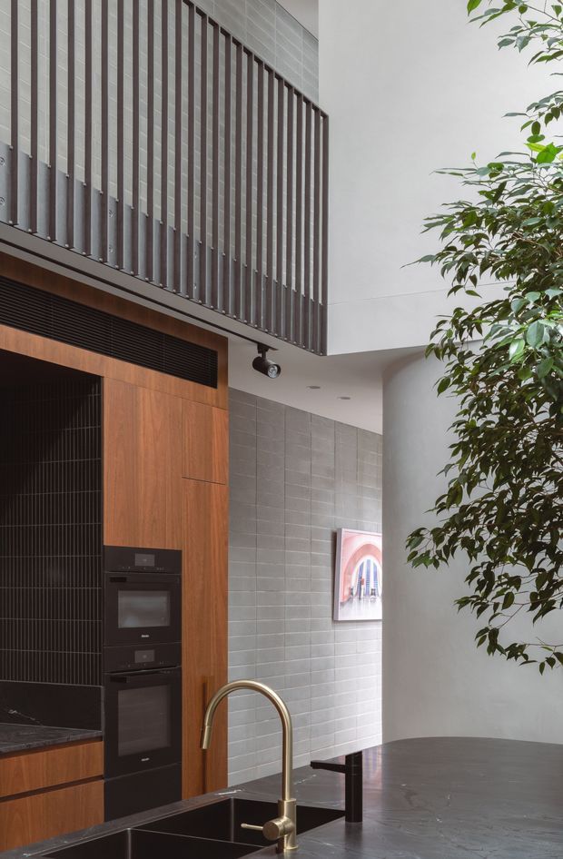 A sophisticated contemporary kitchen viewed from the island, featuring dark stone benchtops, integrated black appliances, warm timber cabinetry, a black vertically ribbed splashback, and a brass-toned gooseneck tap. Pale grey tiled walls and a tall curved plastered wall create a refined architectural backdrop, while vertical dark screening above the kitchen adds texture and height. A leafy indoor tree and framed artwork soften the space.