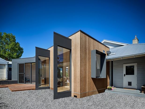 Exterior view of a contemporary residential addition with a steep gabled timber-clad volume, dark metal-framed floor-to-ceiling glazing, and a connected weatherboard-clad house with a low-pitched metal roof. A timber deck and gravel forecourt surround the building.