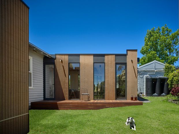 Exterior view of a contemporary timber-clad residential extension connected to an older weatherboard house. The addition has a low, rectilinear form with vertical timber screening, dark-framed full-height glazing, and a raised timber deck opening onto a lawned garden. A small pitched-roof outbuilding is visible to the side.