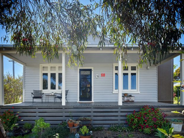 Front exterior view of a compact single-storey weatherboard house with a broad covered veranda, white-painted horizontal cladding, white-framed windows, a dark glazed entry door, and a low horizontal-slat front fence. Mature trees and planted garden beds frame the facade.