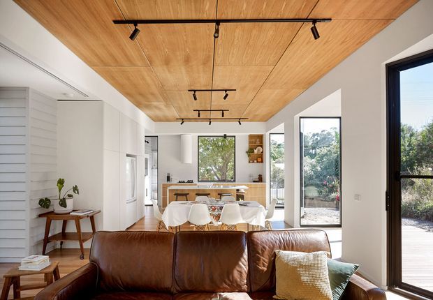 Bright open-plan interior combining a living room, dining area and kitchen. A large brown leather sofa occupies the foreground, facing a dining table with white slipcovered chairs and a compact kitchen beyond. The room features white walls and cabinetry, extensive timber ceiling lining with black track lighting, large windows and glazed doors opening to an outdoor deck or garden. The overall design is warm, contemporary and light-filled.