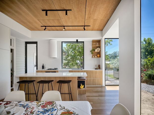 A bright open-plan kitchen and dining area with a timber-lined ceiling, white walls, black-framed glazing, and light timber cabinetry. The kitchen features a long island with seating for three, integrated cooktop, open shelving, and a large window overlooking greenery. A dining table with patterned textiles occupies the foreground, while a glazed door opens to the exterior.