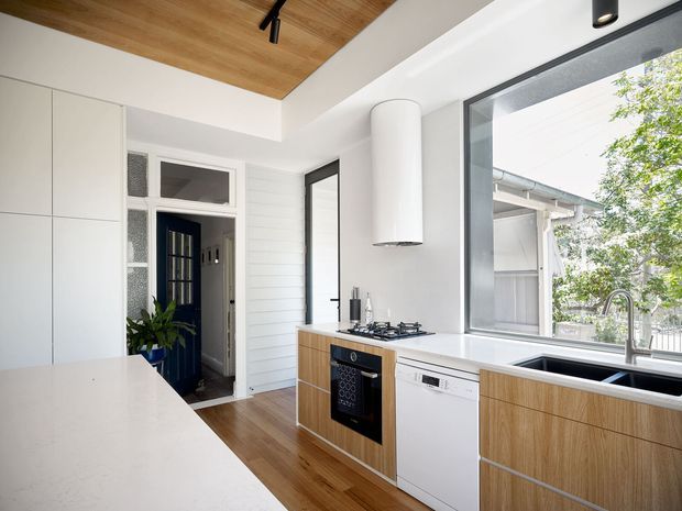 Bright contemporary kitchen with white full-height cabinetry, a large pale stone island, timber-look flooring, and a timber-lined ceiling recess. The kitchen includes a built-in oven, gas cooktop, integrated dishwasher, stainless sink, and light timber drawer fronts. Large black-framed windows provide abundant natural light, while a dark blue glazed door and adjoining hallway are visible beyond the kitchen.
