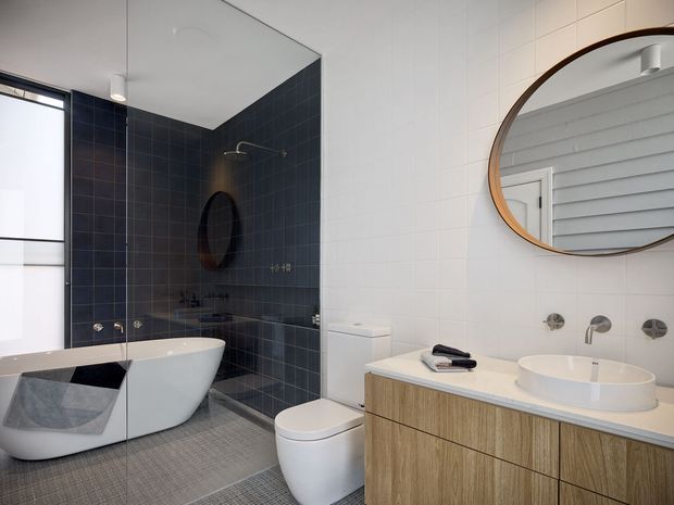 Contemporary bathroom with a freestanding white bathtub positioned beside a dark tiled shower wall, a wall-hung toilet, and a timber-look floating vanity with a white vessel basin. White wall tiles, a large round timber-framed mirror, dark charcoal shower tiles, and patterned grey floor tiles create a crisp, modern contrast. A louvred window is reflected in the mirror.