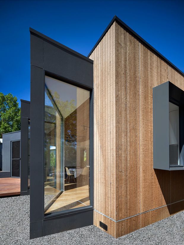 Exterior architectural photograph of a contemporary single-storey home with a sharply pitched gable volume clad in vertical timber boards, contrasted with dark charcoal wall and window framing. Large floor-to-ceiling glazing reveals a dining area inside, while gravel landscaping and a small timber deck surround the building.