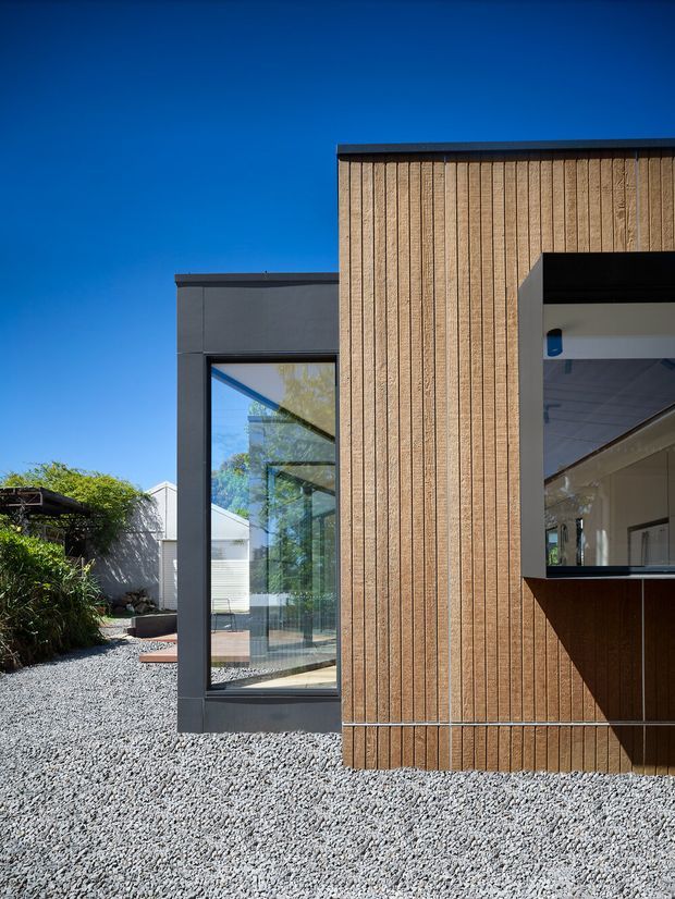 Exterior architectural photograph of a contemporary building composed of a dark charcoal glazed volume and a taller warm timber-clad volume. Vertical timber battens, large black-framed windows, and a recessed opening define the facade. A gravel-covered courtyard and planted garden are visible beside the building under a clear blue sky.