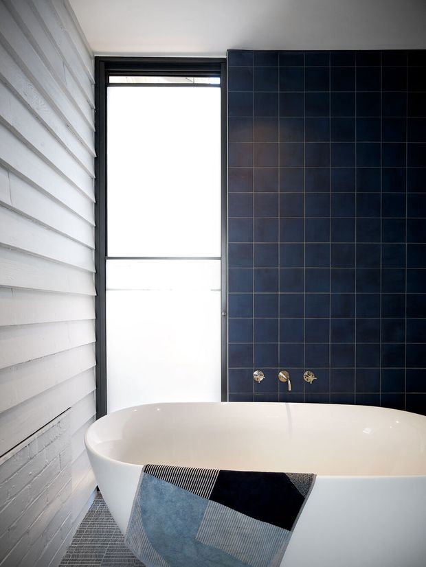 A contemporary bathroom featuring a freestanding white bathtub, a dark navy-blue tiled feature wall with wall-mounted brass tapware, a tall frosted window with a black frame, and white-painted horizontal wall panelling. A geometric blue, black and grey textile is draped over the tub.