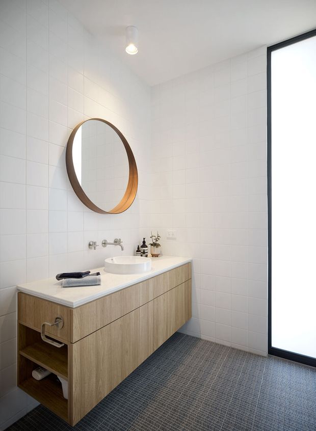 A bright, contemporary bathroom featuring a floating timber-look vanity with drawers and open shelving, a white vessel basin, wall-mounted chrome tapware, and a large round timber-framed mirror. The walls are clad in white square tiles, while the floor has small dark grey textured tiles. A full-height black-framed glazed opening brings in natural light, complemented by a simple pendant light.