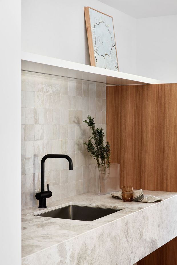 Close-up view of a refined contemporary kitchen sink area. A black gooseneck tap sits above an integrated dark stainless-steel sink set into a pale, heavily veined stone benchtop with a matching waterfall edge. The backsplash is clad in softly textured off-white square tiles, while warm timber cabinetry forms the right side and lower joinery. Minimal styling includes a glass vase with greenery, amber glassware, and a framed artwork above the cabinetry.