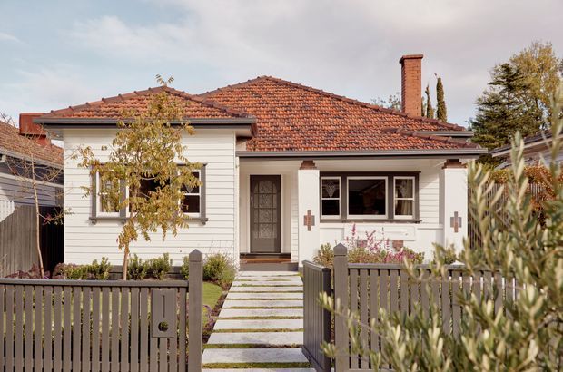 Front exterior view of a single-storey weatherboard cottage with a low-pitched terracotta tiled hipped roof, brick chimney, traditional timber-framed windows, and a small covered porch. A paved stepping-stone path leads through a dark picket fence to the entry, with established planting around the frontage.