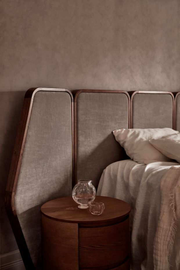 A warm, intimate bedroom detail featuring a tall upholstered headboard with curved timber framing, layered cream bedding and pillows, and a round dark timber bedside table with a glass carafe and tumbler. The room has a textured taupe plaster wall, soft directional lighting, and a refined earthy palette.