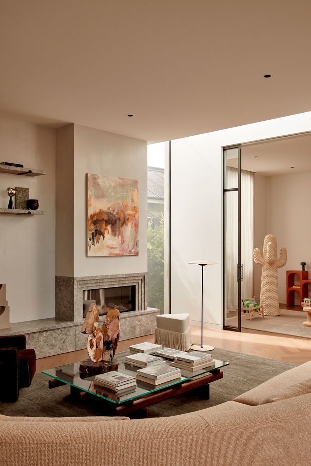Warm, refined contemporary living room with a large textured sectional sofa, glass-topped coffee table, sculptural objects, abstract artwork, and a substantial stone fireplace. Full-height glazing and an open glass doorway connect to a bright adjoining space, while a play area and sculptural cactus decor are visible beyond. The room combines neutral plaster walls, timber flooring, stone fireplace cladding, and carefully curated furnishings.