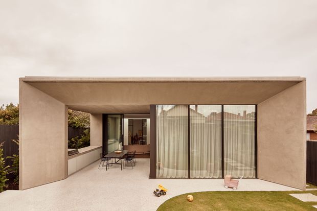 A minimalist single-storey residential facade with a deep recessed covered terrace or courtyard. Smooth warm beige rendered walls frame large dark-framed floor-to-ceiling glazing with sheer curtains, opening onto a pale paved outdoor area and a small lawn. The flat roof and strong rectangular geometry create a calm, contemporary composition.