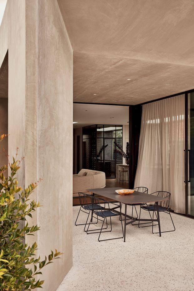 A refined contemporary dining and living area with a long dark dining table and black wire-frame chairs set on a pale terrazzo floor. A textured plaster or stone-like feature wall and matching warm-toned ceiling create a monolithic backdrop, while full-height black-framed glazing with sheer curtains opens the room to natural light. A partial sofa, indoor planting, and a glimpse of a kitchen or hallway are visible beyond the dining area.