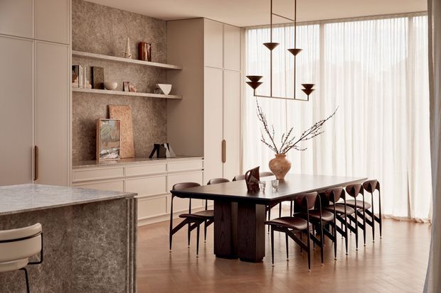 Warm, refined open-plan dining area with a long dark timber dining table and matching chairs, positioned beside a stone-finished kitchen island. Floor-to-ceiling sheer curtains provide soft natural light, while built-in cream cabinetry and open shelving form a feature wall. A sculptural multi-pendant chandelier hangs above the table, complemented by neutral decorative objects and a vase of branches.