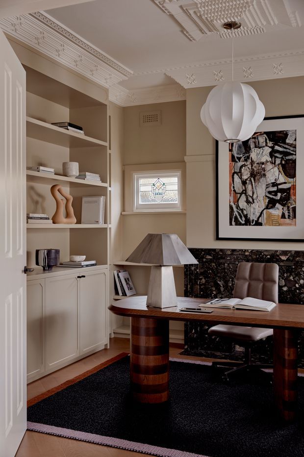 A refined home office with warm taupe walls, ornate decorative plaster ceiling and cornice detailing, built-in shelving and cabinetry, a dark stone fireplace surround, and a substantial dark timber desk. A sculptural white pendant light, geometric table lamp, leather office chair, abstract artwork, books, and a dark rug complete the richly detailed interior.