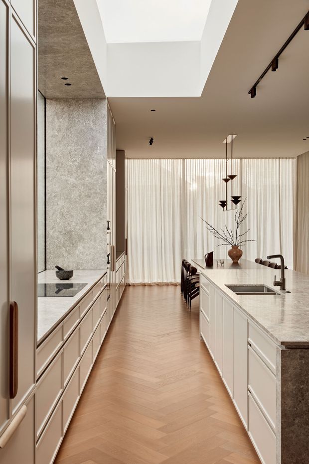 A refined open-plan kitchen and dining area viewed along a central circulation aisle. The kitchen features pale framed cabinetry on both sides, a large stone island with an integrated sink and dark tapware, and a tall stone-clad wall with open shelving. Warm timber herringbone flooring leads to a dining table positioned in front of full-width sheer curtains. A large ceiling opening or skylight brings in natural light, while sculptural pendant lights hang above the dining area. The space has a calm, warm, contemporary classic character with restrained neutral finishes.